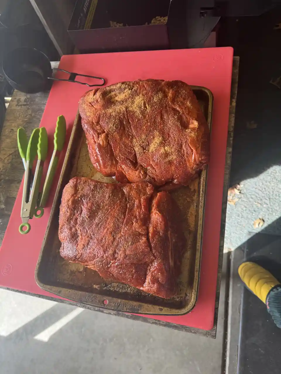 Two large, heavily seasoned raw meat cuts sit on a metal baking sheet placed on a red cutting board. Beside the tray are…