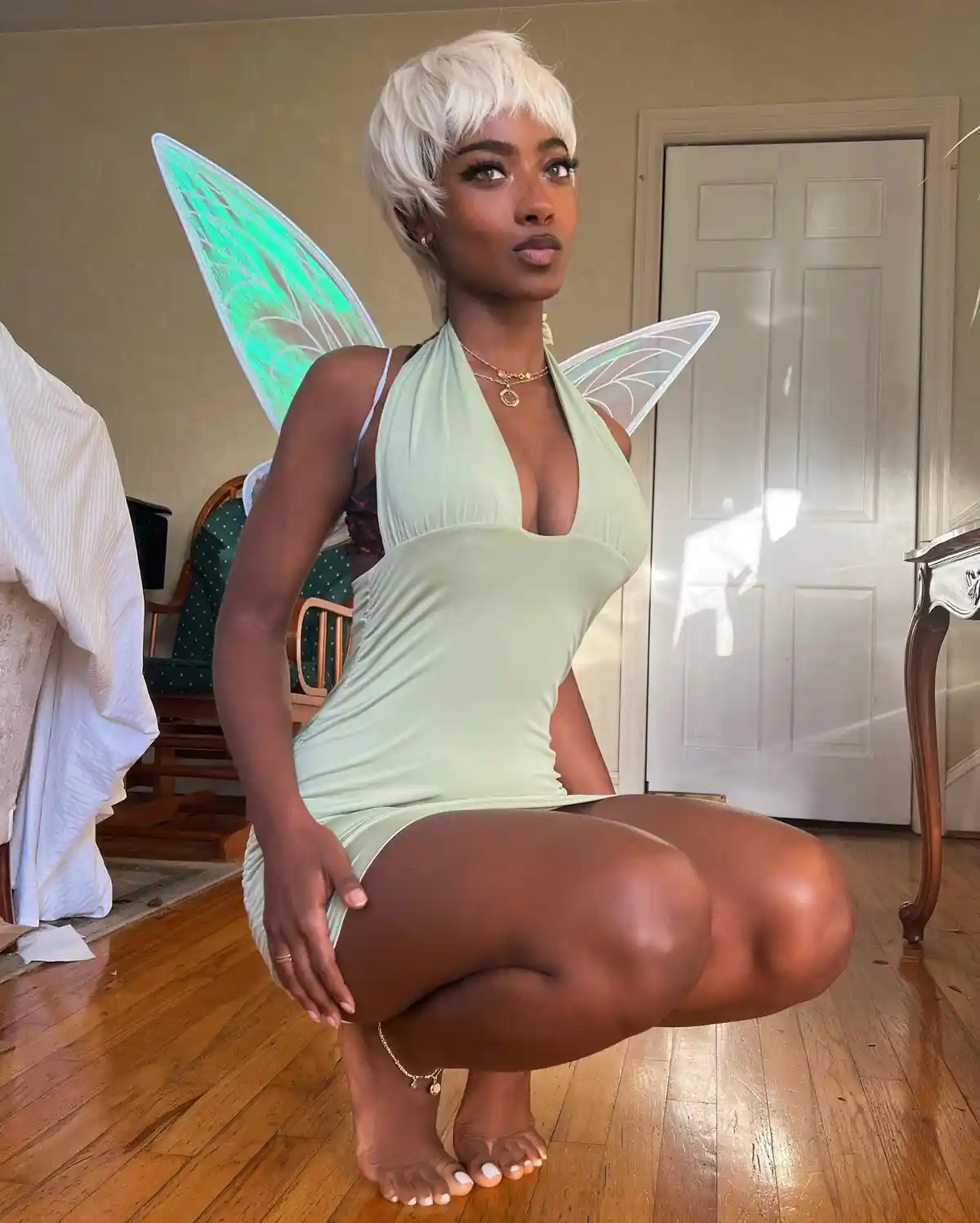 A woman with short white hair and glowing green fairy wings crouches on a wooden floor in a light green halter dress…