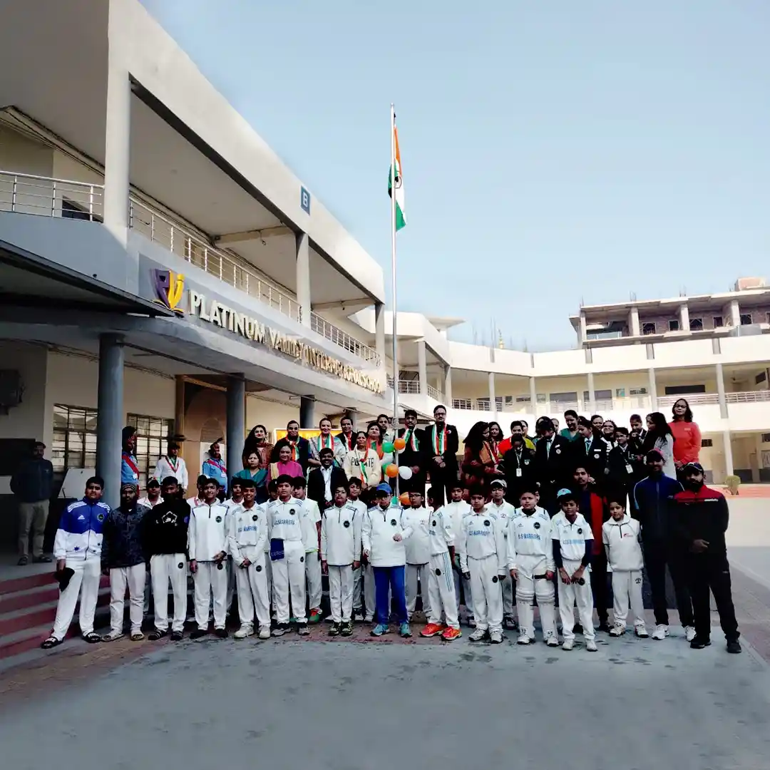 A group photo of students and staff in front of the Platinum Valley International School building, with the Indian flag…