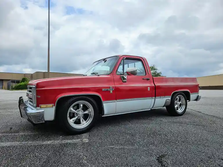 A red and silver 1980s Chevrolet pickup truck with chrome wheels is parked in an asphalt lot under a cloudy sky. The text…