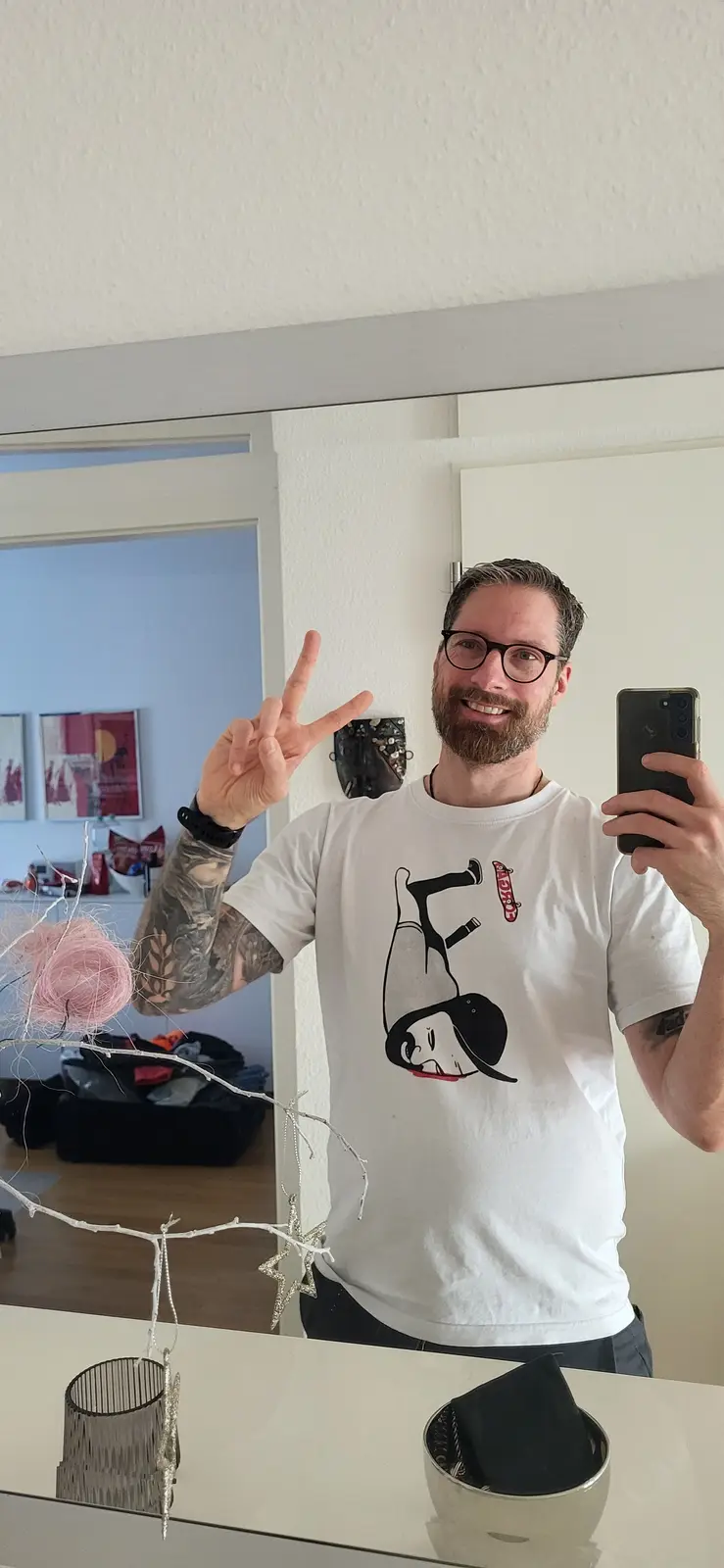 A man with glasses and a beard takes a mirror selfie while holding a phone in one hand and making a peace sign with the…