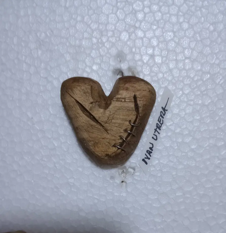 A wooden heart-shaped object with a visible crack and stitched repair sits on white bubble wrap. A small white tag next to…