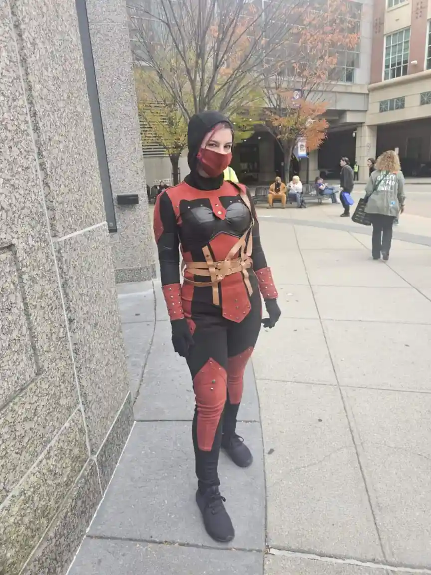 A person in a detailed red and black assassin costume with a hood and face mask stands on a city sidewalk. The costume…