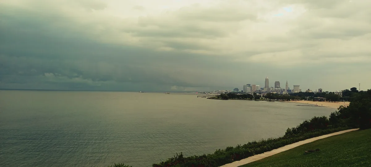 A wide, moody photo of a city skyline across a large body of water under heavy clouds, with a grassy foreground and path…