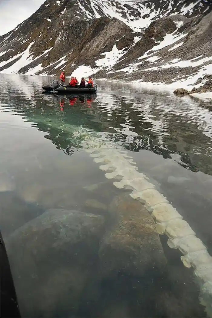 A group of people in orange life jackets are in a small inflatable boat on a calm, clear lake surrounded by snow-dusted…