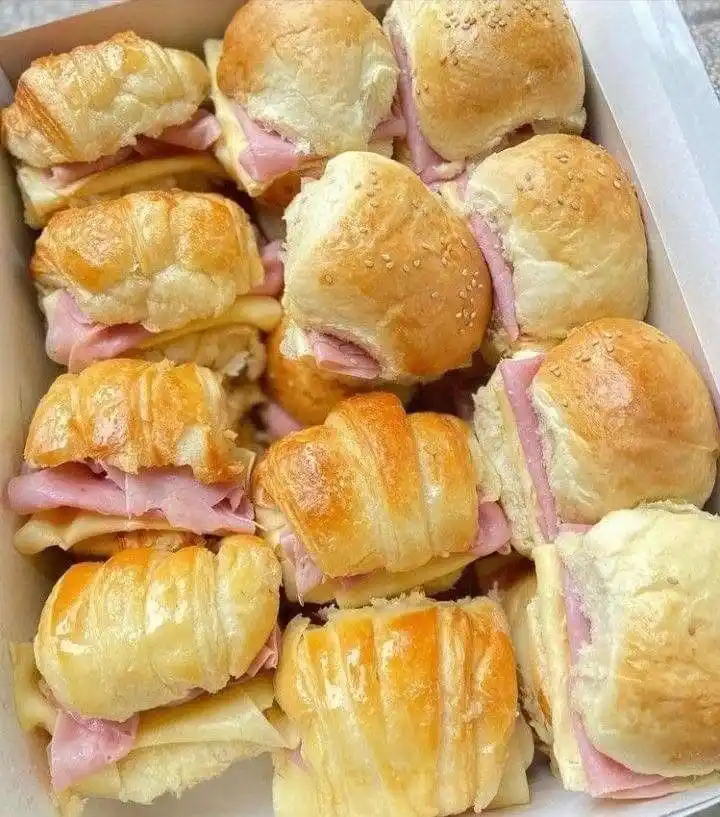 A white cardboard tray is filled with various types of small sandwiches, including ham and cheese sliders and…