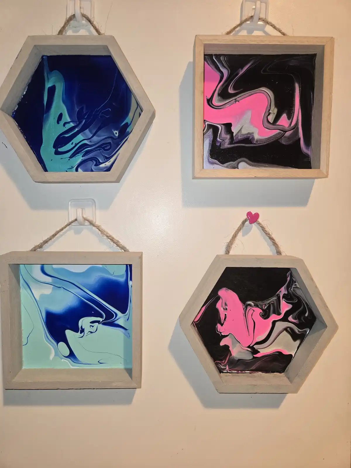 Four small framed artworks hang on a wall, two hexagonal and two square, each containing swirling abstract paint in vibrant…