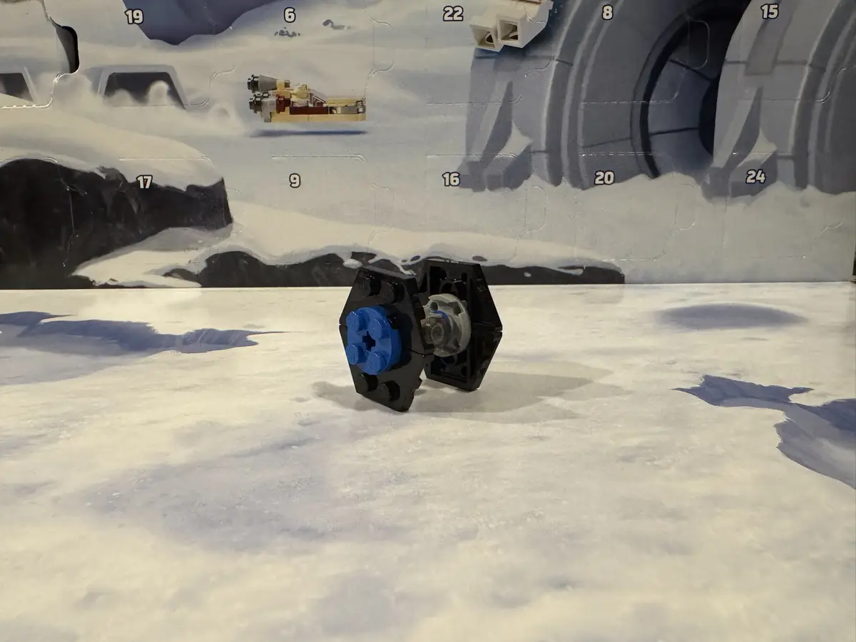 A single, partially assembled Lego piece, featuring a blue stud and black connector, sits on a snowy surface in front of a…