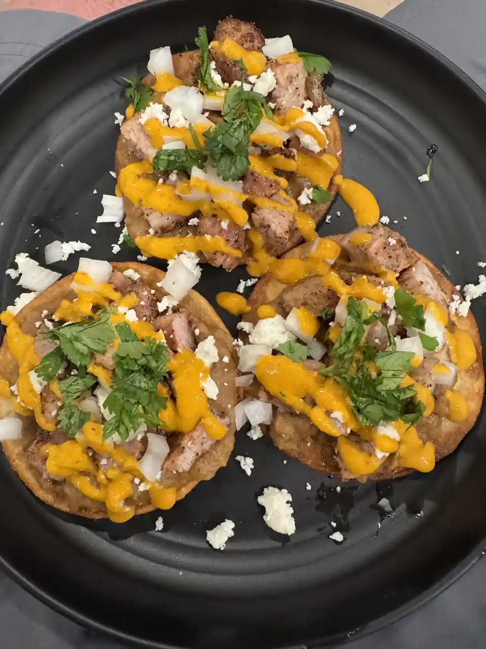 Three tostadas on a black plate, topped with meat, yellow sauce, chopped white onions, and fresh cilantro. The image has…