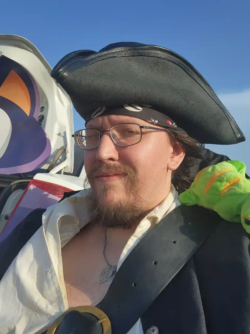 A man dressed as a pirate with a tricorn hat, bandana, glasses, and a white shirt with a black vest stands outdoors next to…