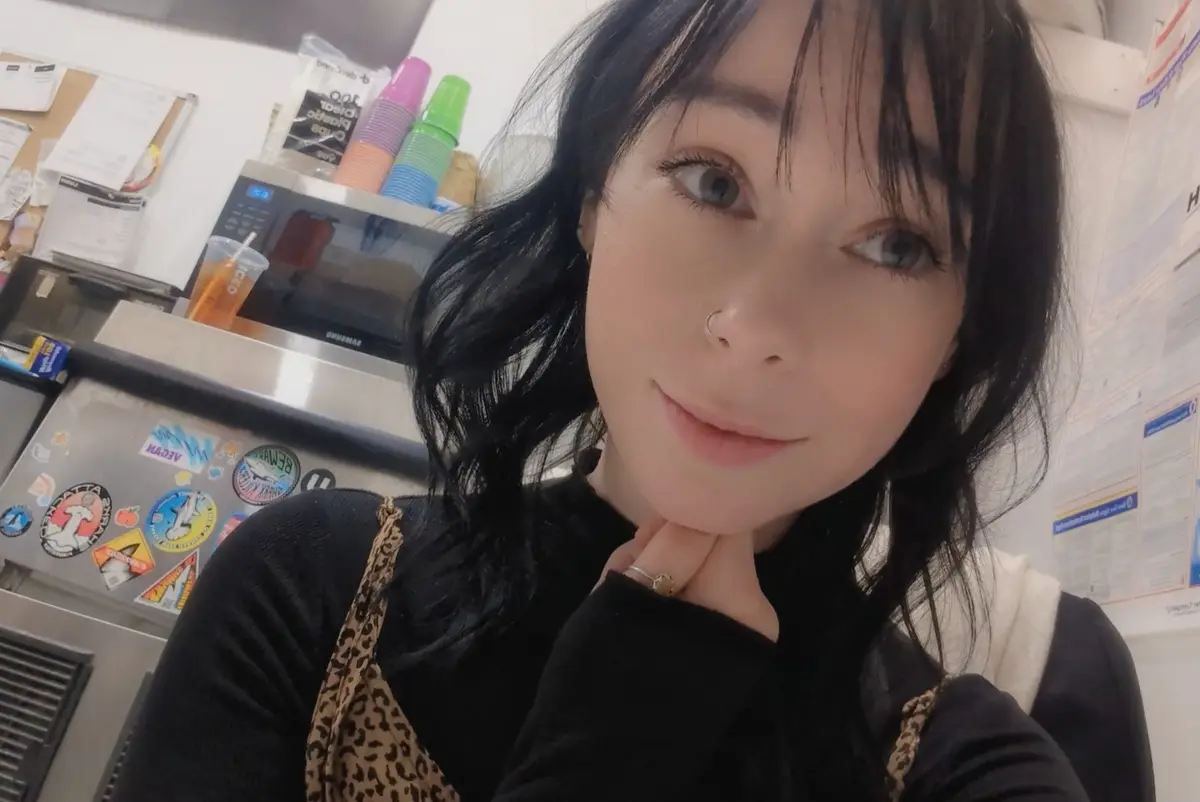 A young woman with dark hair and a nose ring takes a selfie in what appears to be a kitchen or cafe, resting her chin on her…