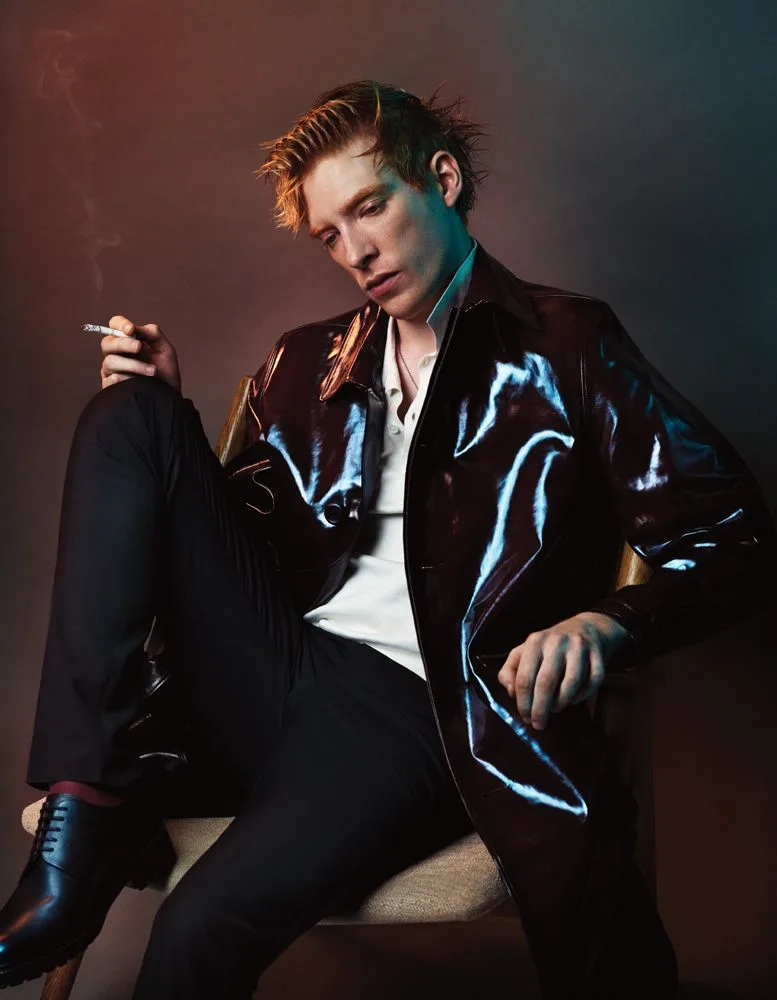 a man with spiky hair sits in a chair wearing a shiny dark jacket over a white shirt and black pants, holding a cigarette…