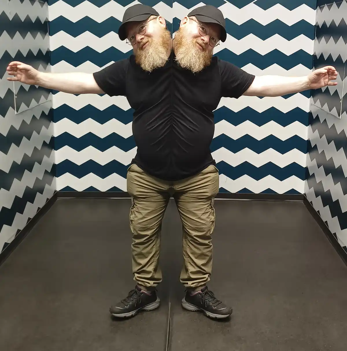 A person with a beard, glasses, and a black cap stands in front of a zigzag-patterned wall, arms outstretched, creating a…