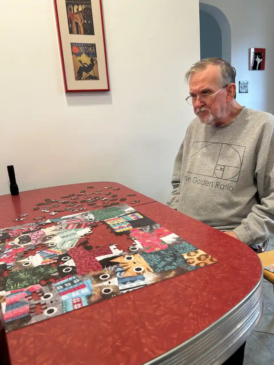 An older man with glasses and a gray sweatshirt featuring a Fibonacci spiral and the text 'the Golden Ratio' sits at a red…