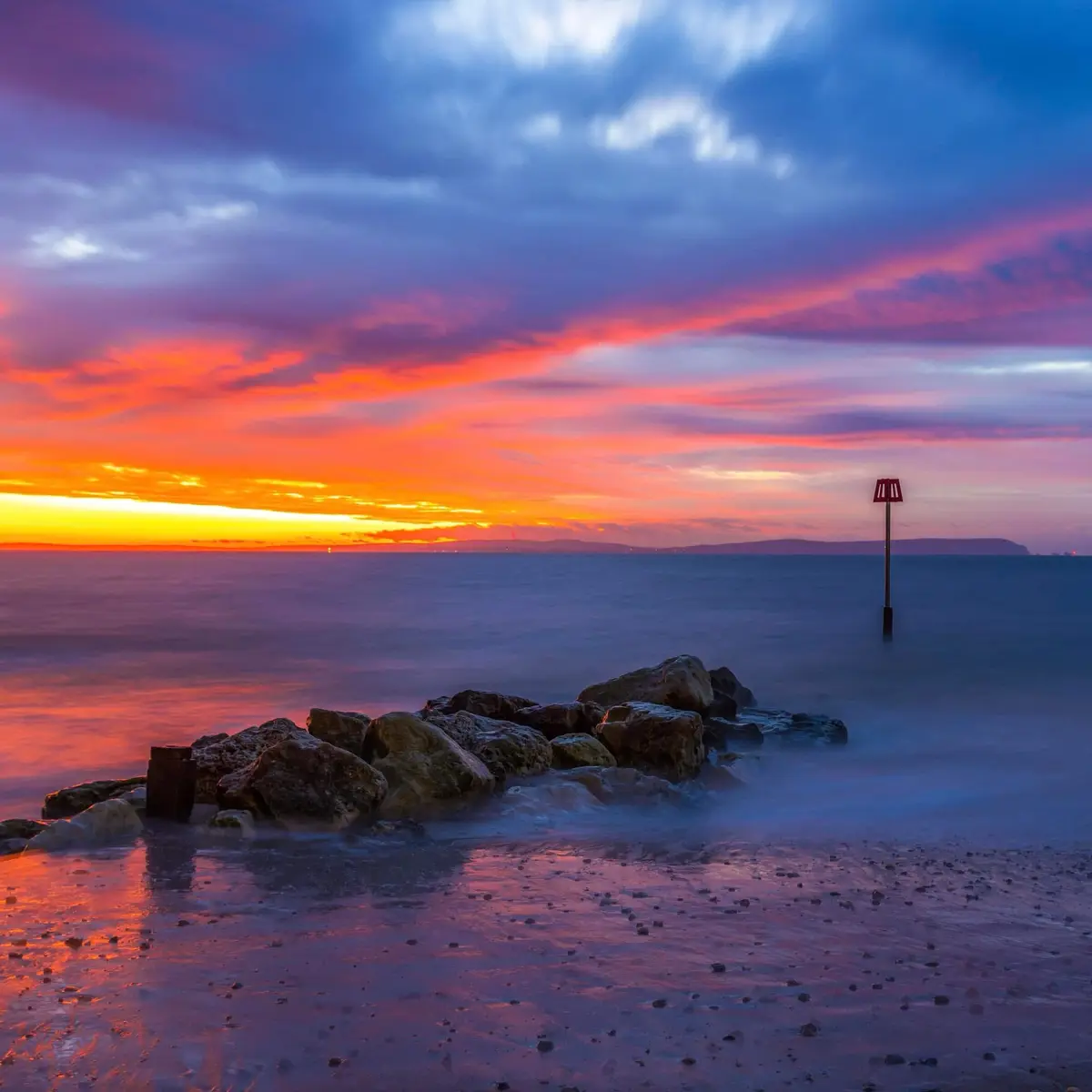 A long-exposure photograph of a serene beach at sunset, featuring vibrant orange, pink, and purple clouds reflected in calm…