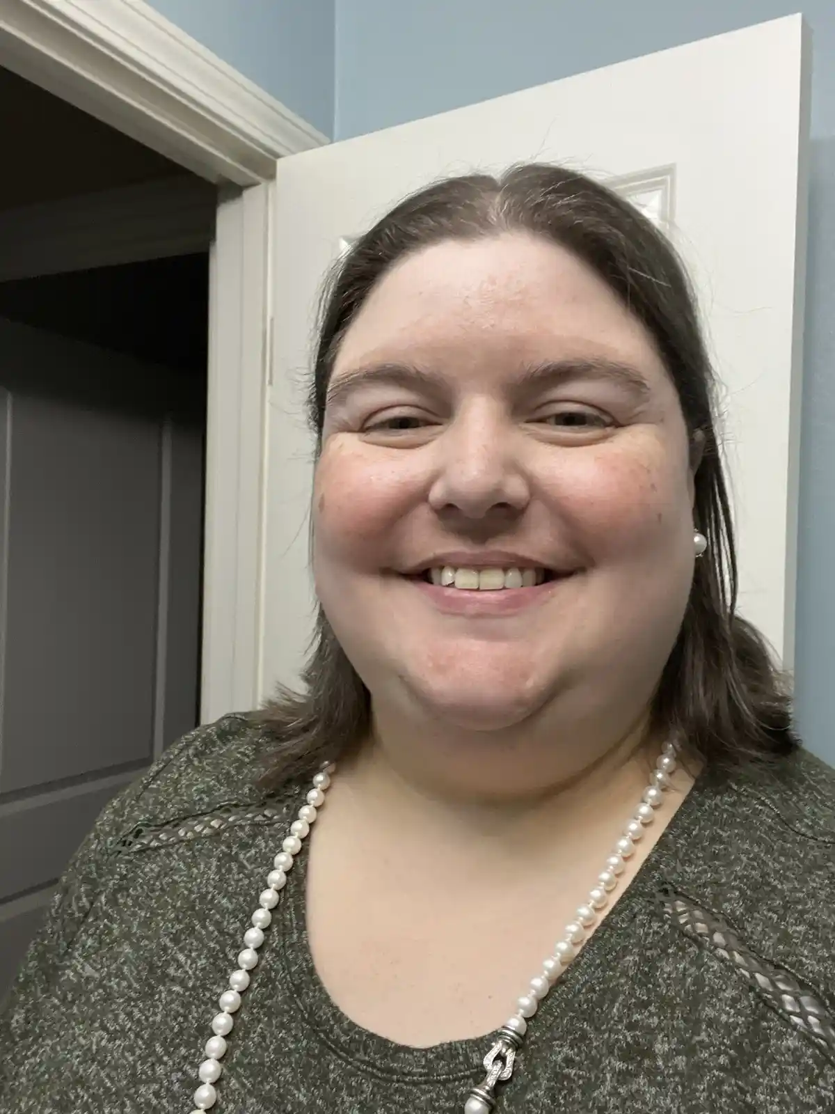 A smiling woman with brown hair, wearing a green top and a pearl necklace, stands in front of a white door with a light blue…