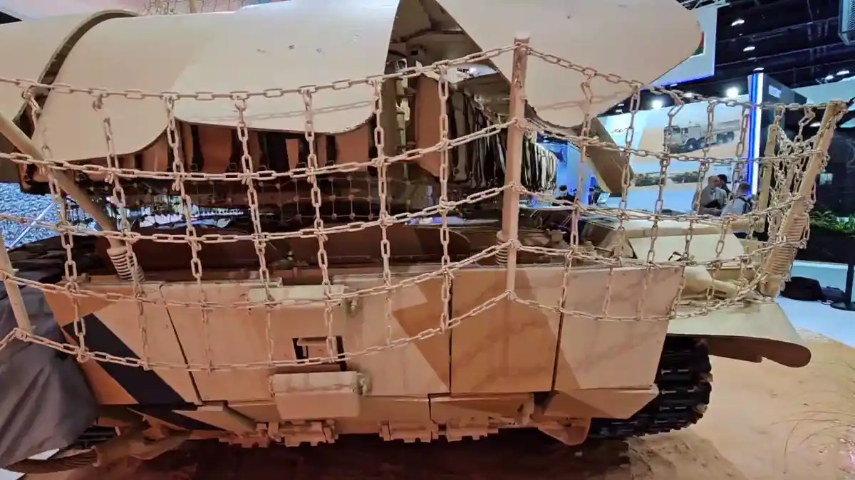 a tan-colored military tank is shown from the side, with a chain-link netting structure covering its upper hull and turret…