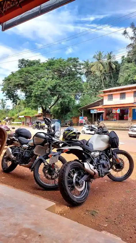 Two adventure-style motorcycles are parked side-by-side on a dirt patch under a bright blue sky with palm trees and…