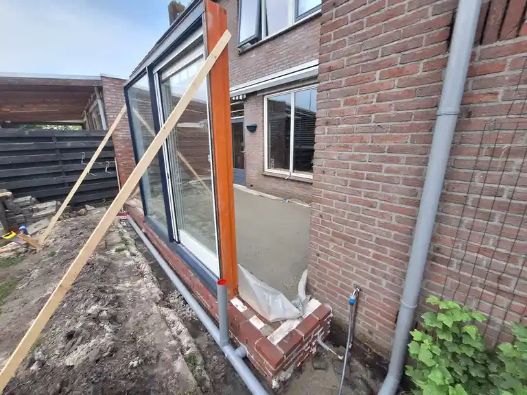 A large glass window frame is being installed on a brick house, propped up by wooden braces and surrounded by construction…