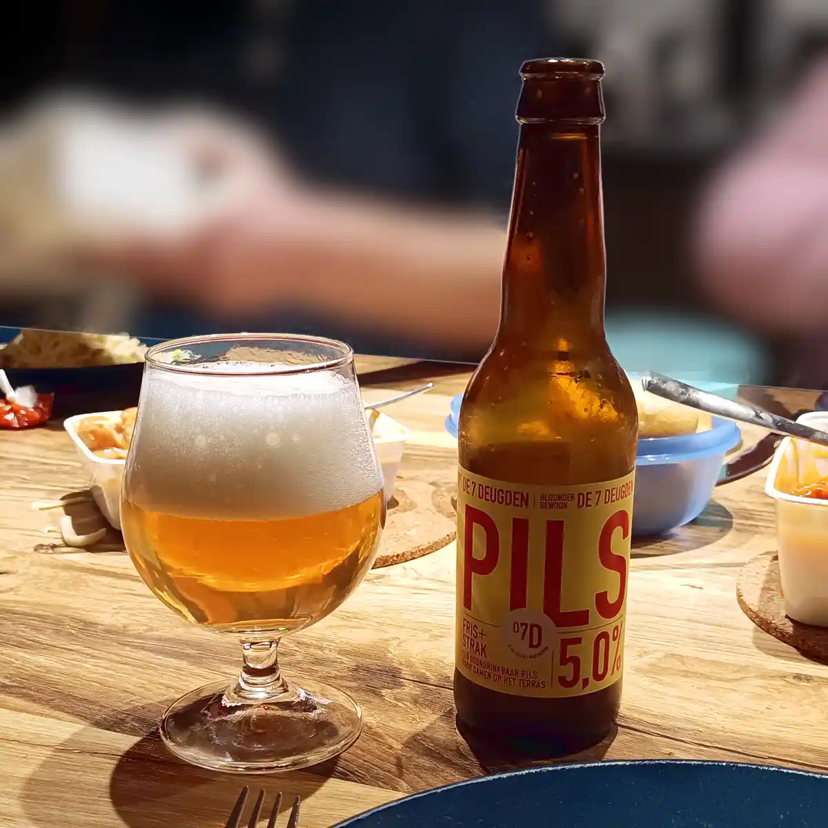 A chilled bottle of 'Pils 5.0%' beer from 'De 7 Deugden' sits next to a frothy glass on a wooden table, surrounded by food…