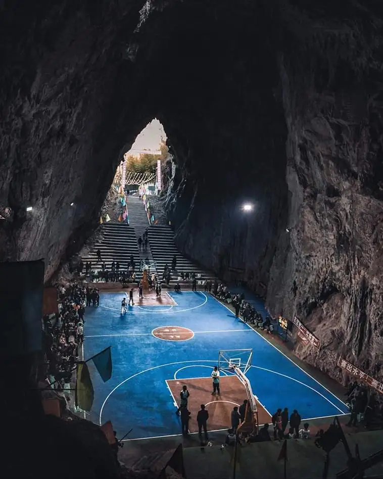 A wide-angle view of a blue basketball court set inside a large, naturally formed cave with rough stone walls. People are…