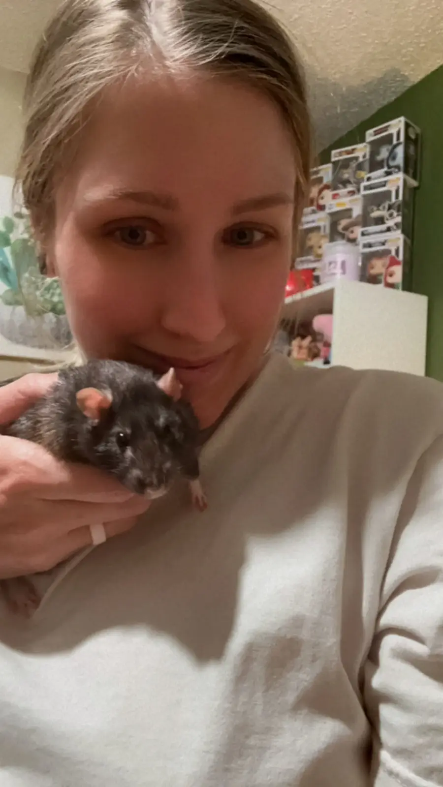 A person with light brown hair and a white sweatshirt holds a small black rat close to their face, smiling slightly. The rat…