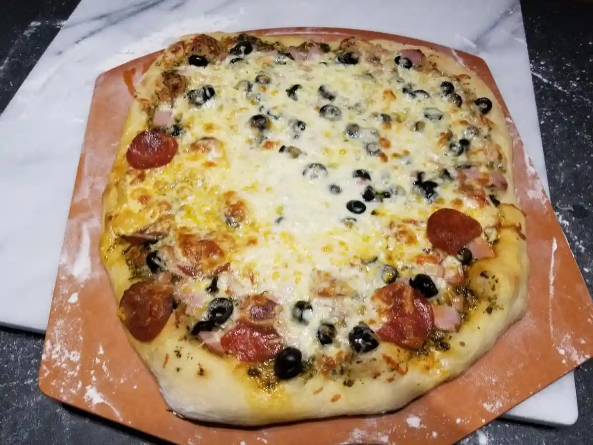 A homemade rectangular pizza with pepperoni, black olives, and ham sits on a floured orange pizza stone atop a marble…