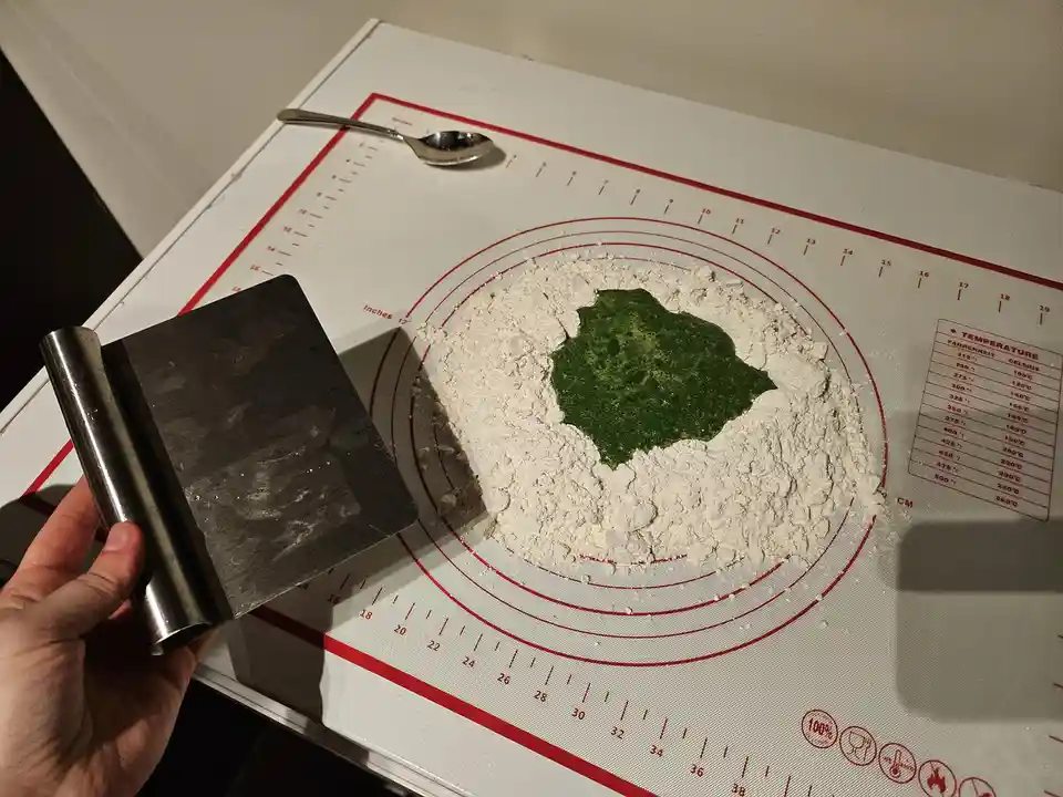 A hand holds a metal dough scraper over a white silicone baking mat marked with red measurement lines and temperature…