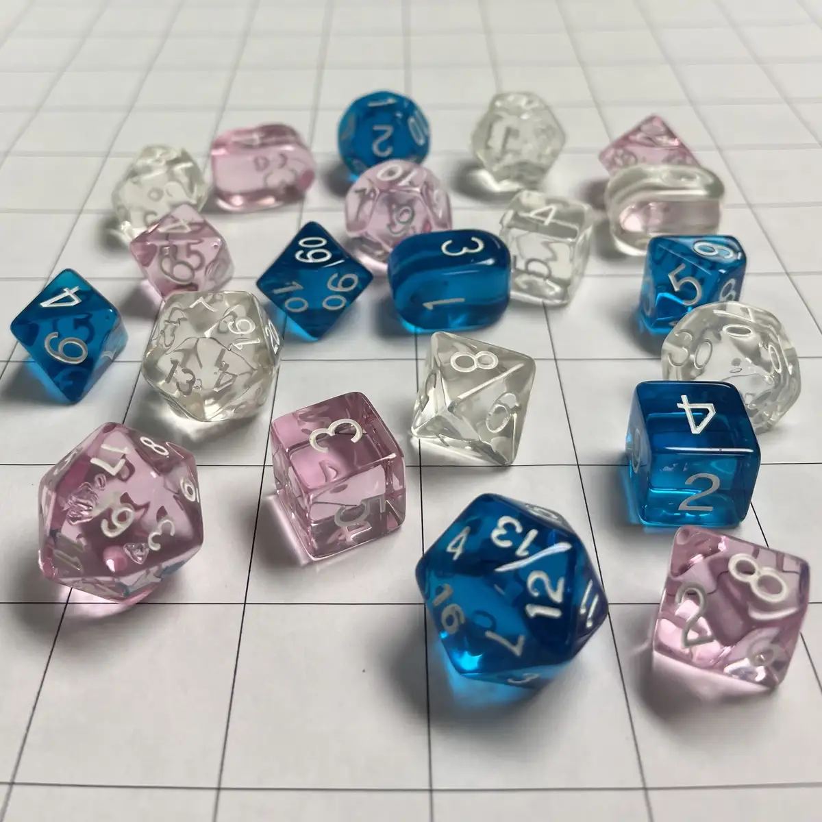 A collection of colorful, translucent polyhedral dice in blue and pink, scattered on a white gridded surface, with various…