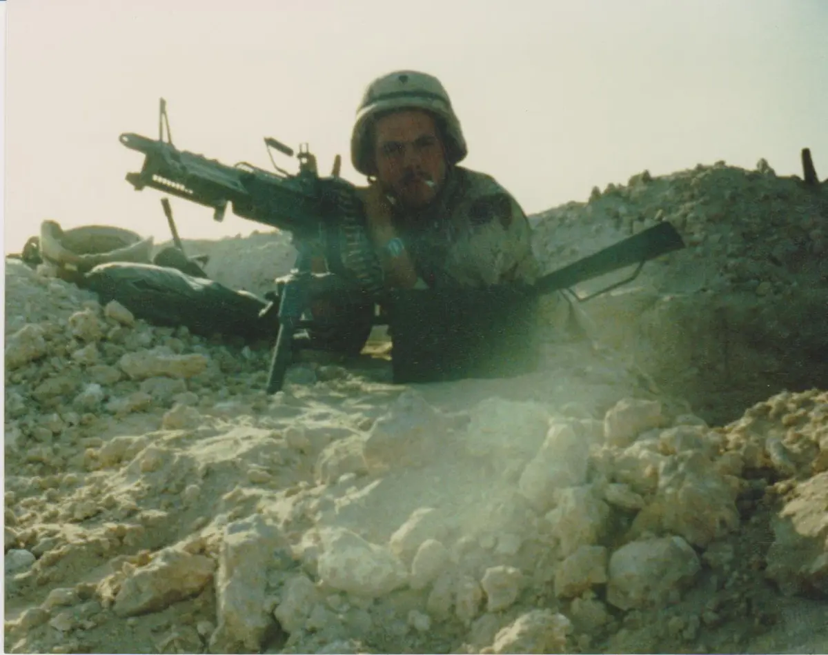 a soldier in a helmet and camouflage is lying prone in a rocky, dusty terrain, holding a heavy machine gun. He has a…