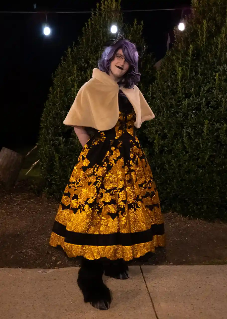 A person in a full demon queen costume stands outdoors at night, wearing a black and gold floral dress, a beige cape, purple…