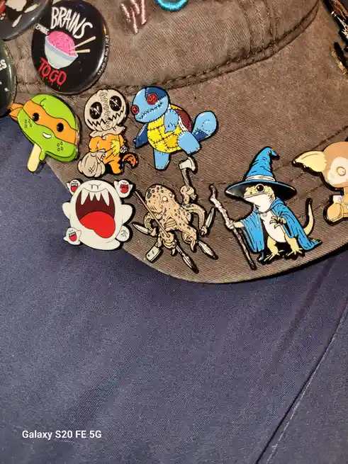 A close-up photo shows a brown cap covered in various colorful enamel pins, including a green ninja turtle, a stitched doll…
