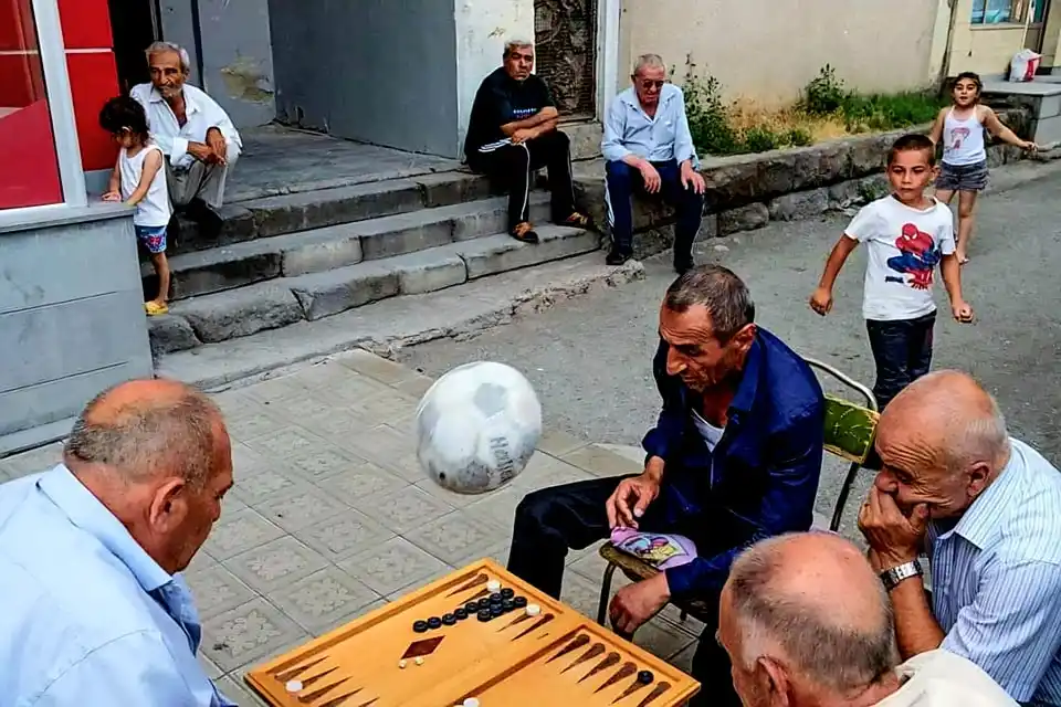 A group of men are gathered around a backgammon board outdoors, seemingly engrossed in a game. A soccer ball is suspended…