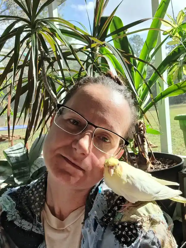 A person wearing glasses and a patchwork jacket smiles at the camera while a yellow and white cockatiel perches on their…