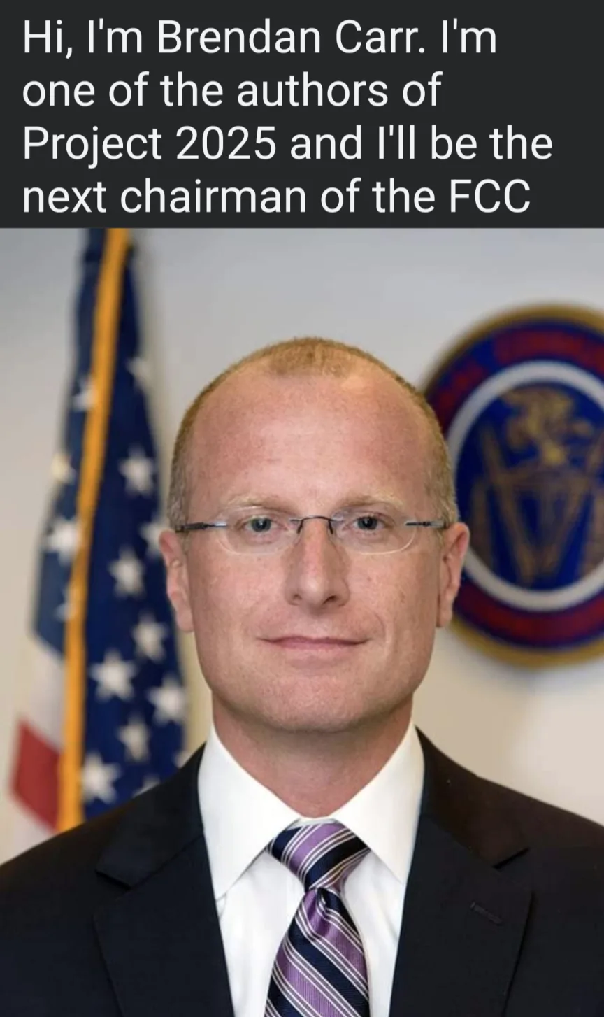 A formal headshot of a man with glasses, wearing a dark suit, white shirt, and striped tie, standing in front of an American…