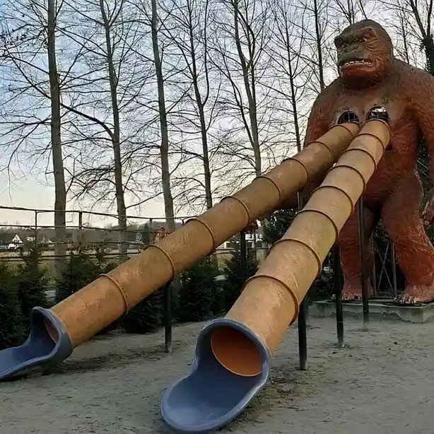 A large, brown gorilla statue stands in a playground with two long, rusty metal slides emerging from its chest, ending in…