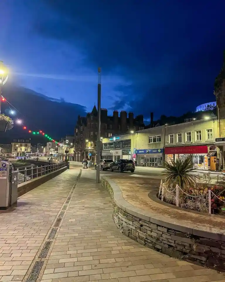 A nighttime photo of a quiet, well-lit town street with shops, a paved walkway, and festive string lights against a dark…