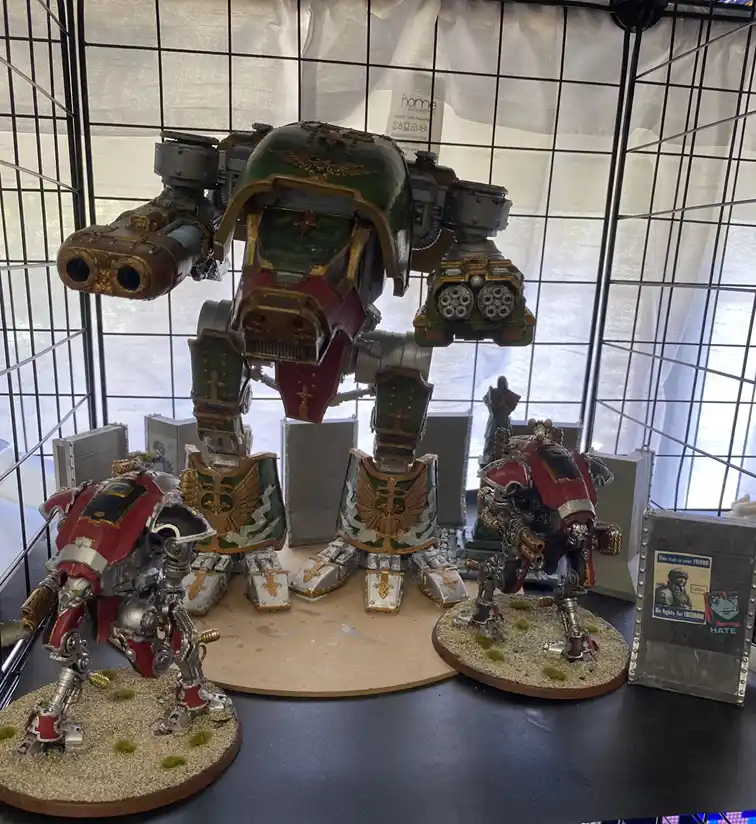 A large, heavily armored green and gold war machine stands center stage, flanked by two smaller red and silver robotic…