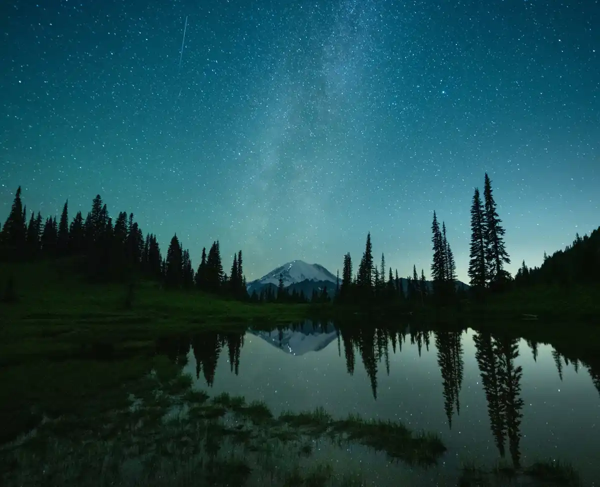 A serene night photograph shows a snow-capped mountain, likely Mount Rainier, framed by dark pine trees and reflected in a…