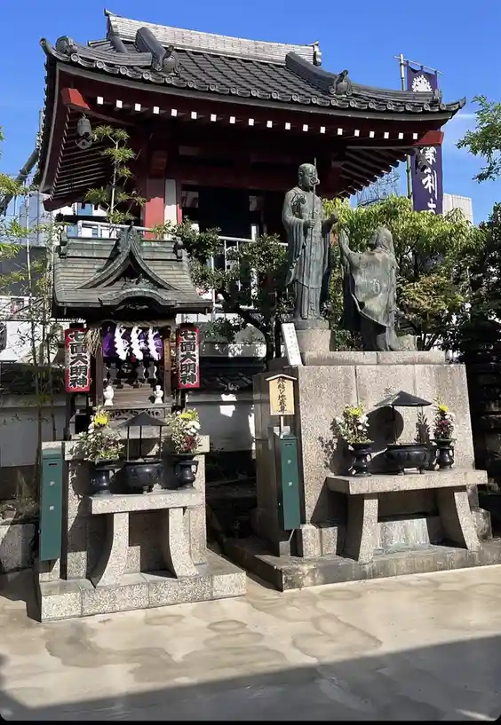 An outdoor shrine with two bronze statues of figures, one standing and one kneeling, set against a traditional Japanese…