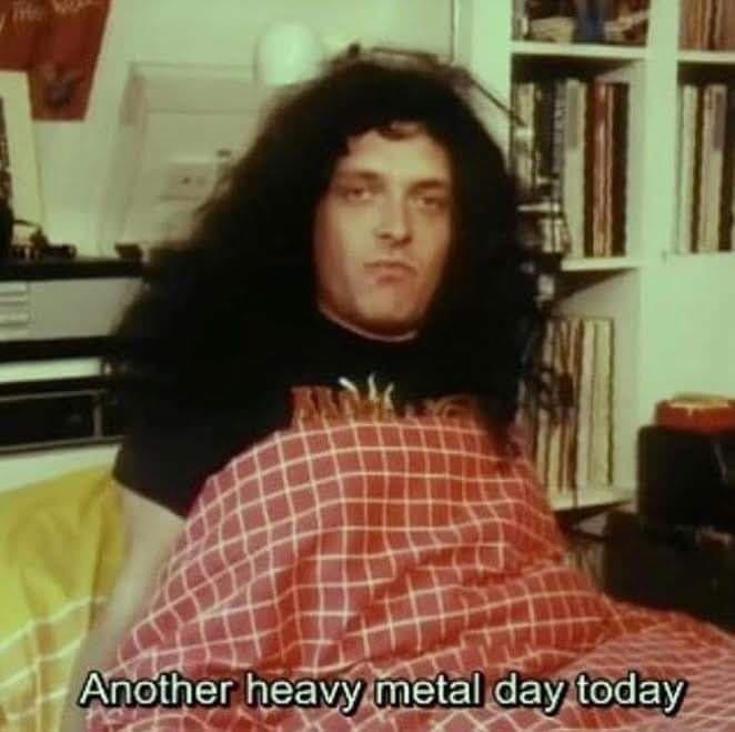 A man with long, dark, wavy hair sits in a room, covered by a red and white checkered blanket. Behind him, a bookshelf…