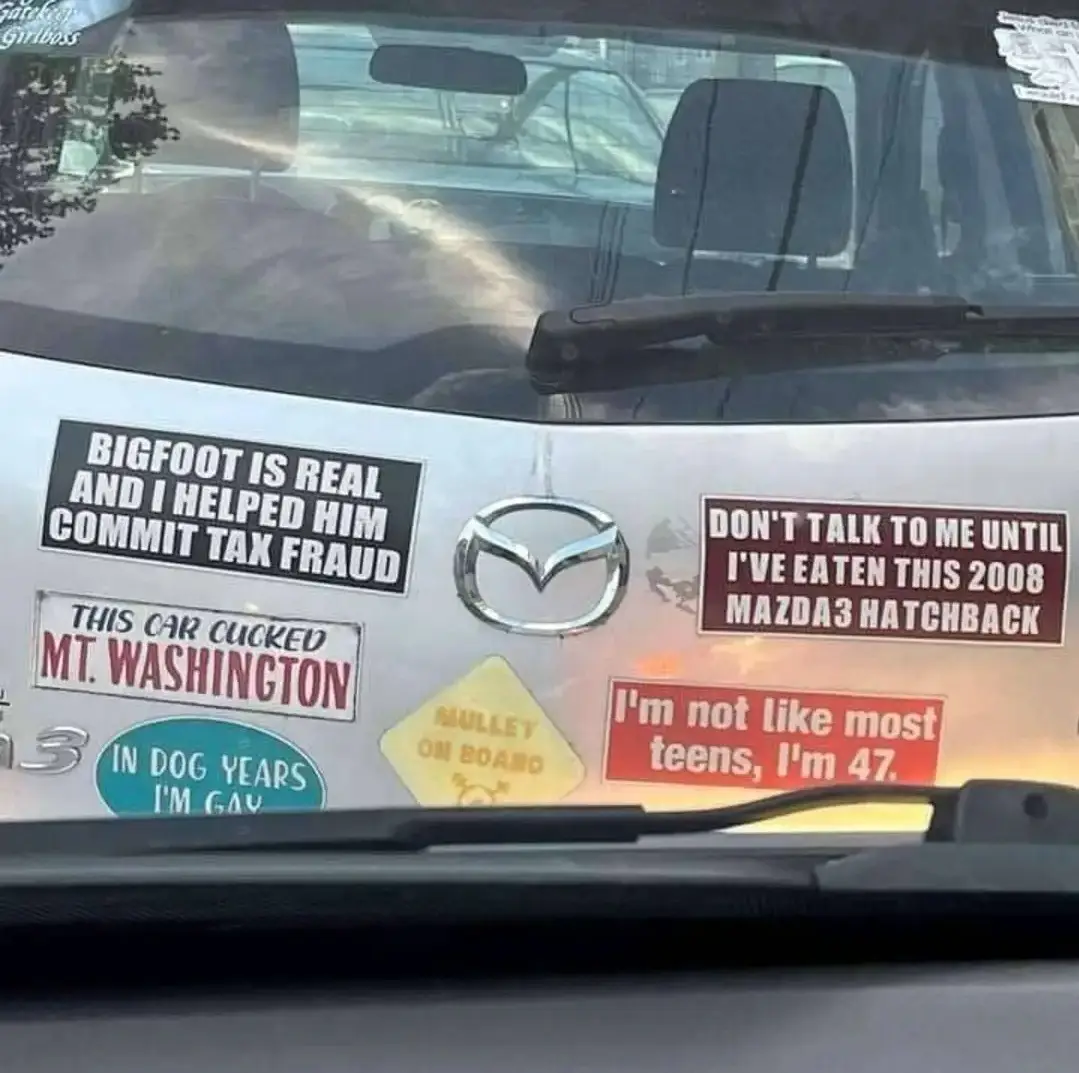 The image shows the rear of a silver 2008 Mazda3 hatchback covered in humorous car decals. Visible stickers include 'BIGFOOT…