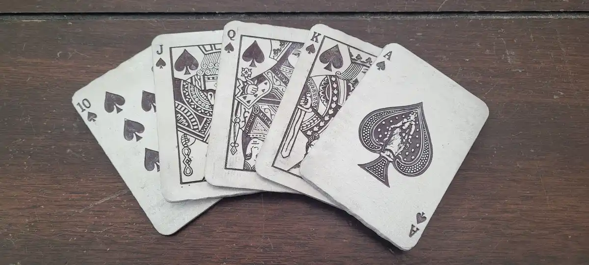 Five playing cards, all spades, fanned out on a dark wooden surface, showing the 10, Jack, Queen, King, and Ace. The…