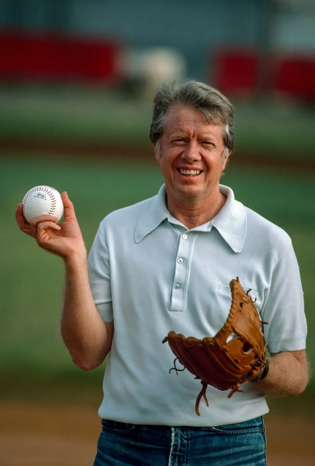 A smiling man with gray hair, wearing a light blue polo shirt and jeans, stands on a baseball field holding a baseball in…