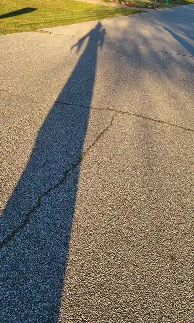 A long, dark shadow of a person walking on a cracked asphalt driveway is cast by low-angle sunlight, making the shadow…