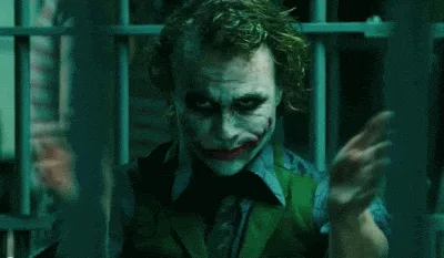 The image shows the character Joker from the movie 'The Dark Knight' in a prison cell, with bars visible behind him. He is…