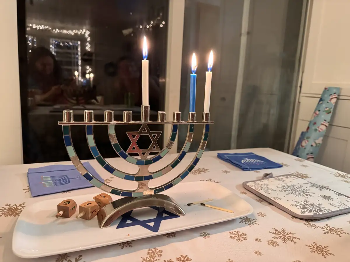 A silver menorah with three lit candles (one white, two blue) sits on a white plate with snowflake-patterned tablecloth…
