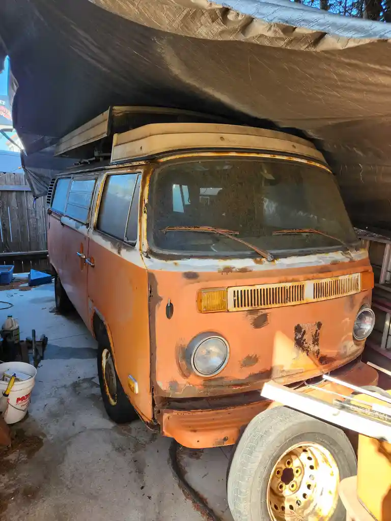 A rusty, orange vintage Volkswagen van with a pop-top roof is parked under a tarp, surrounded by tools and a bucket, visibly…