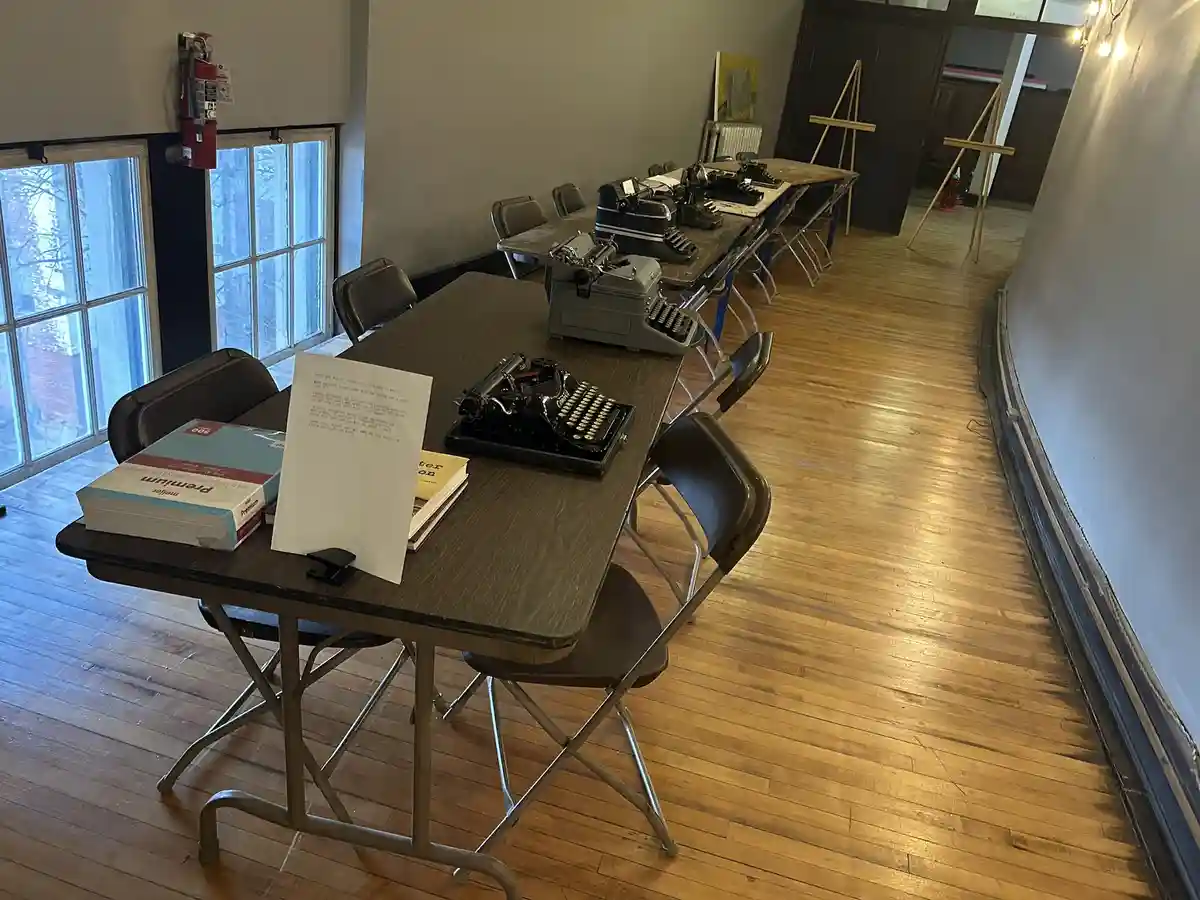 A room with long wooden tables lined with vintage typewriters and folding chairs. On the nearest table, a printed sign with…