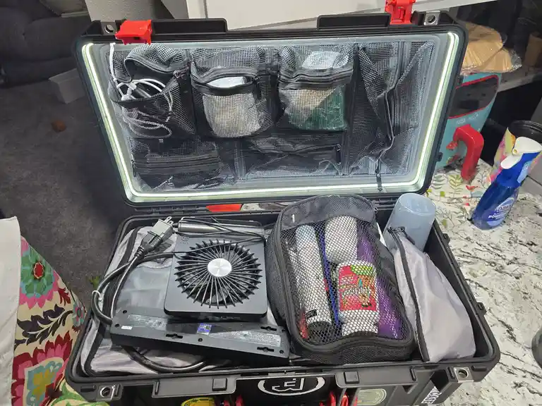 An open black Pelican-style hard case with LED lighting around the lid, filled with organized gear including a portable fan…